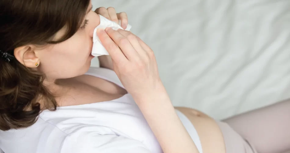 White pregnant woman blocking nose bleeding with tissue while sitting on the bed